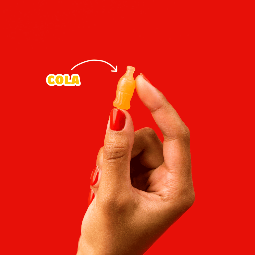 Hand holding a bottle-shaped cola gummy on a red background with an arrow pointing to text: COLA.